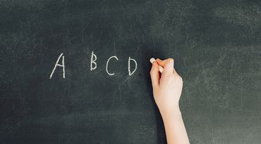 child&#39;s hand with chalk write letters a, b, c, d on black chalkboard
